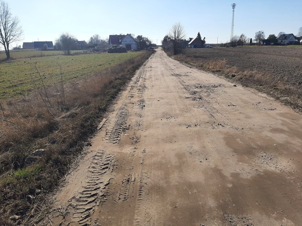 Obraz przedstawia drogę gruntową z dziurami. Droga prowadzi wzdłuż pól w stronę zabudowań.